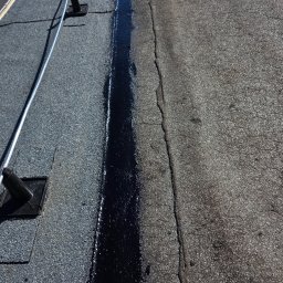 Paweł Porębski Natalia Kalczuk - Uszczelnianie pęknięcia na papowym dachu smołą. Widoczny fragment dachu przed i po naprawie, z elementami mocującymi. Praca dekarska w słoneczny dzień.