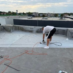 Paweł Porębski Natalia Kalczuk - Pracownik w trakcie naprawy płaskiego dachu budynku komercyjnego. Widoczne narzędzia, kabel zasilający i fragmenty membrany dachowej. W tle zabudowa miejska.