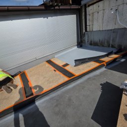 Paweł Porębski Natalia Kalczuk - Prace dekarskie na dachu: montaż izolacji z użyciem taśm i płyt OSB. Widoczny fragment dachu z blachy falistej i ściana budynku. Pracownik w kamizelce odblaskowej.