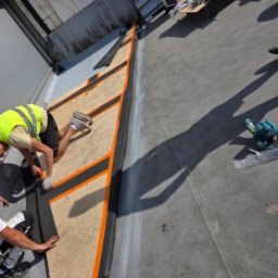 Paweł Porębski Natalia Kalczuk - Montaż izolacji dachu płaskiego z użyciem płyt izolacyjnych i papy termozgrzewalnej. Widoczni pracownicy w trakcie pracy, narzędzia i materiały budowlane.