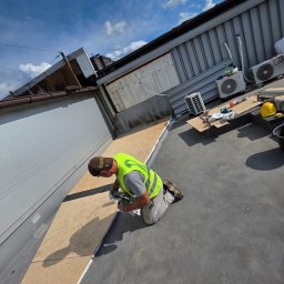 Paweł Porębski Natalia Kalczuk - Pracownik w kamizelce montuje płytę na dachu przemysłowym. Widoczne narzędzia, klimatyzatory i fragment nieba. Prace dekarskie w toku, z bliska.