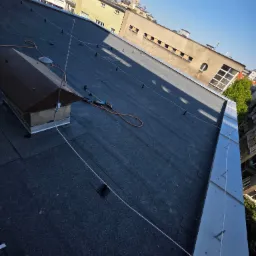 Wykończony dach pokryty papą z zamontowanymi zabezpieczeniami przeciwśniegowymi i obróbką blacharską. Widok z góry na tle zabudowy miejskiej w słoneczny dzień.