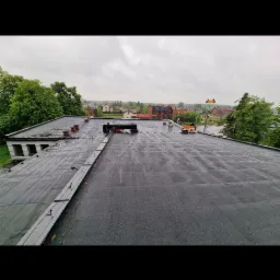 Widok z góry na płaski dach pokryty papą, z widocznymi kominami wentylacyjnymi i elementami mocującymi, w tle zabudowa miejska i zieleń drzew pod pochmurnym niebem.