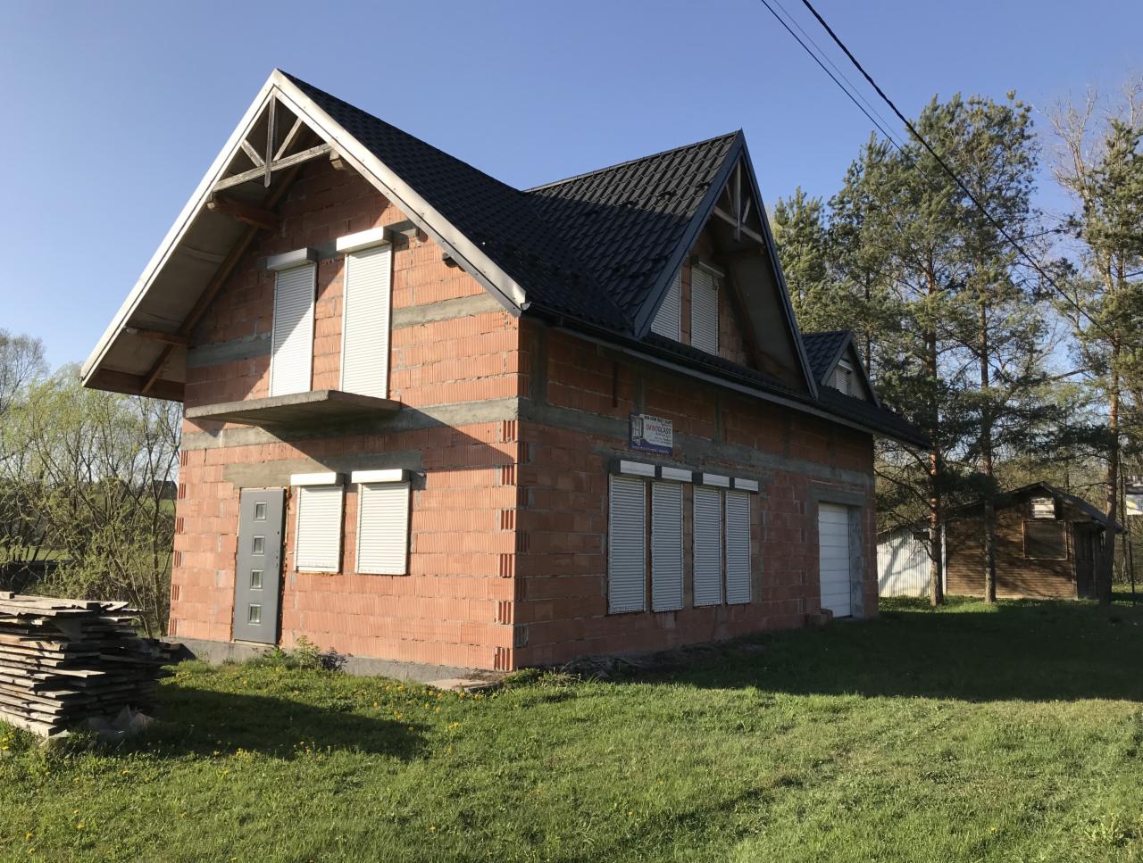 Surowy budynek z czerwonej cegły w trakcie budowy, z zainstalowanymi roletami okiennymi, widoczne deski na trawie i fragment lasu w tle.