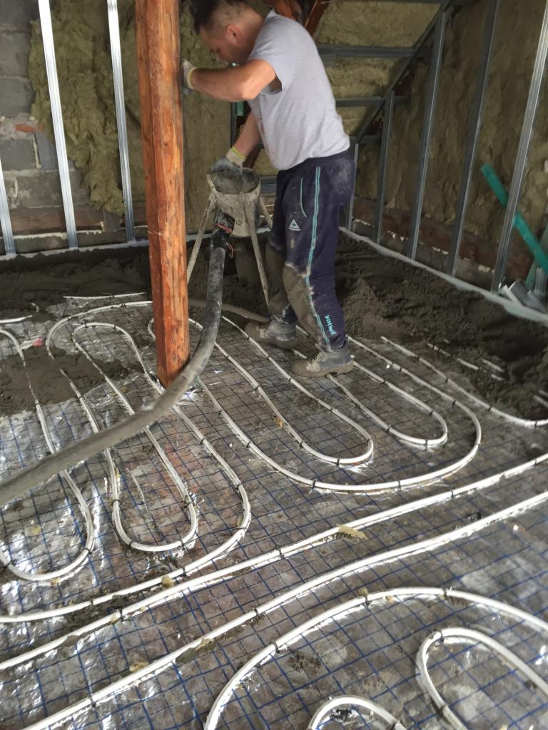 Pracownik w trakcie wylewania betonu na instalację ogrzewania podłogowego w pomieszczeniu z ocieplonymi ścianami i metalową konstrukcją.