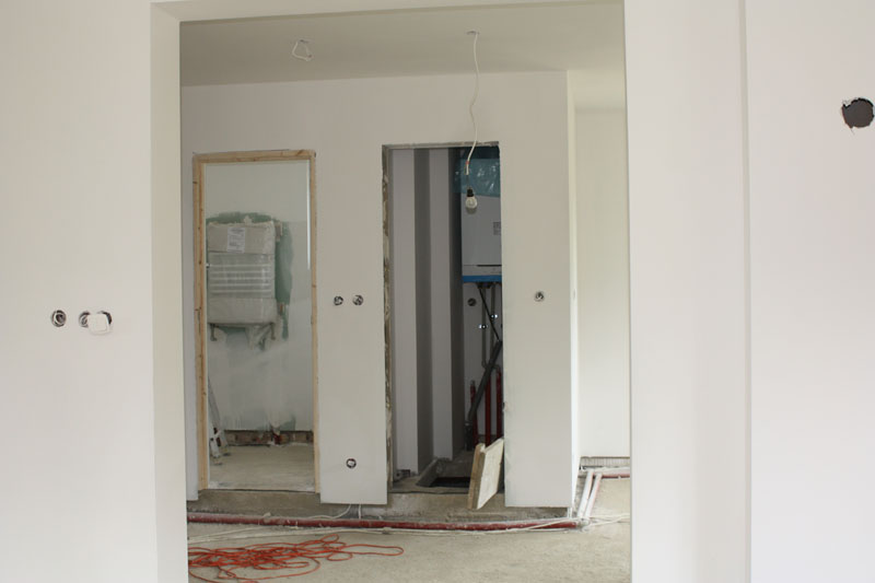 Wnętrze w trakcie wykończenia: białe ściany z przygotowanymi punktami elektrycznymi, widoczne otwory drzwiowe i instalacje grzewcze, betonowa podłoga z rozłożonymi przewodami.