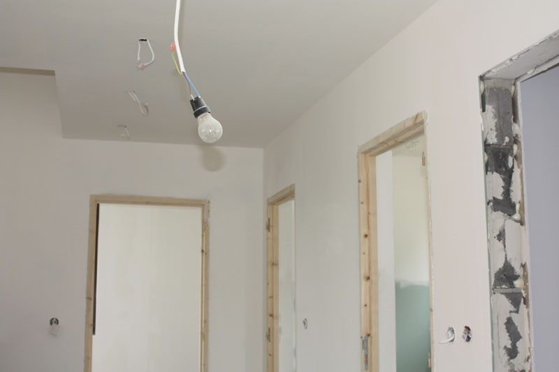 Wnętrze w stanie deweloperskim z surowymi ościeżnicami, świeżo otynkowanymi ścianami i wiszącą żarówką na kablu.