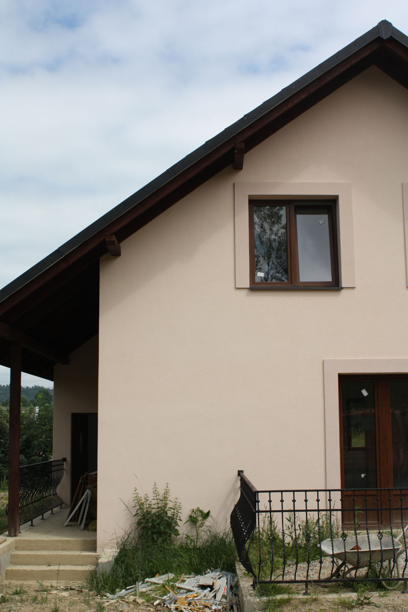 Fragment domu z beżową elewacją, widoczne okno z brązową ramą, fragment zadaszonego tarasu z metalową balustradą oraz nieuporządkowany teren wokół budynku.