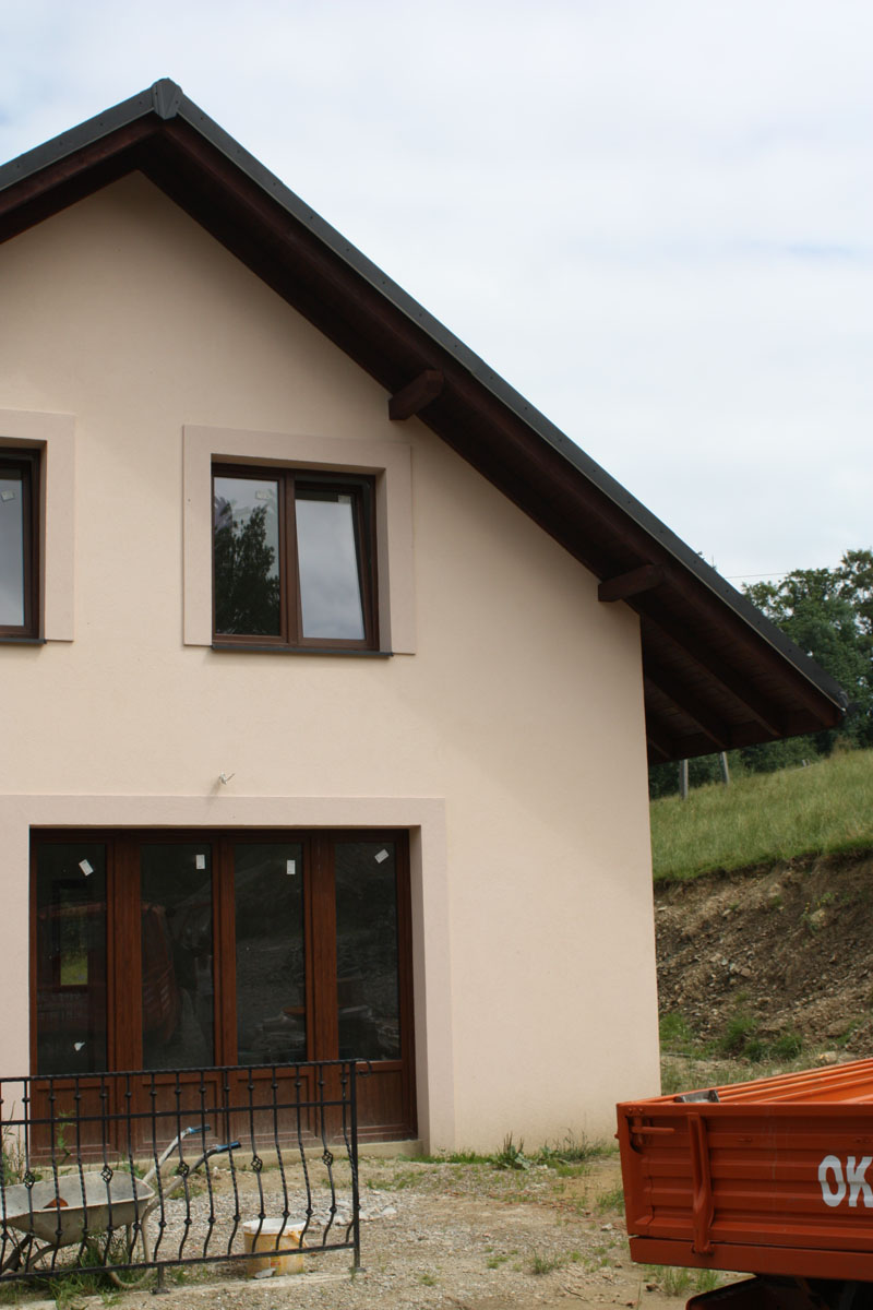 Nowo otynkowana elewacja domu z brązowymi oknami i drzwiami, widoczna przyczepka samochodowa i taczka na tle zieleni.