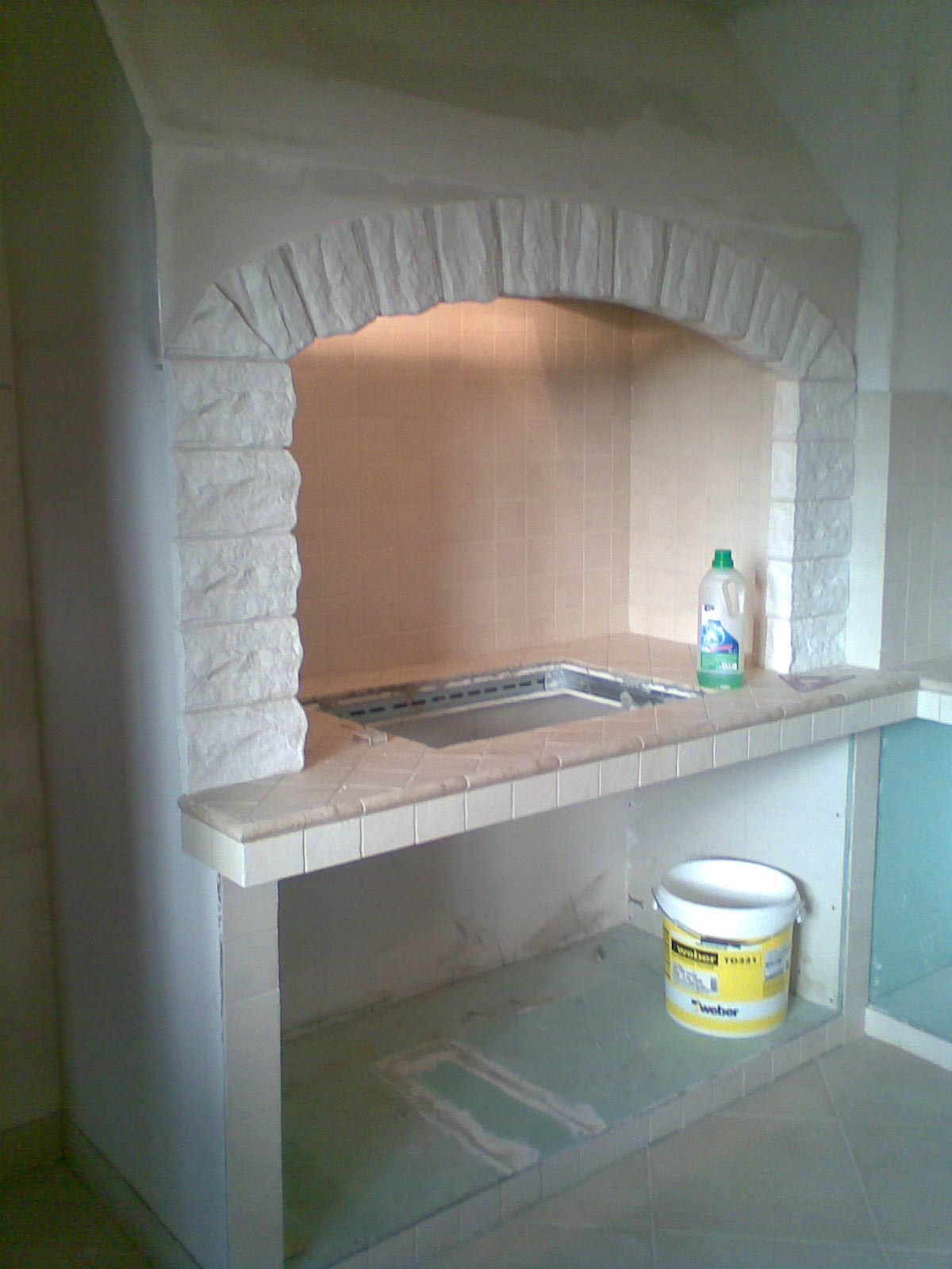 Wnętrze wnęki z kamienną obudową łukową, wyłożonej płytkami, z widocznym zlewem i wiadrem z zaprawą budowlaną, w trakcie prac wykończeniowych.