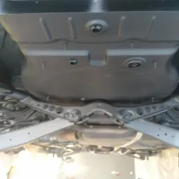 Podwozie samochodu po zabezpieczeniu antykorozyjnym, widok od spodu, metalowe elementy zawieszenia i płyta podłogowa pokryte czarną, matową powłoką.