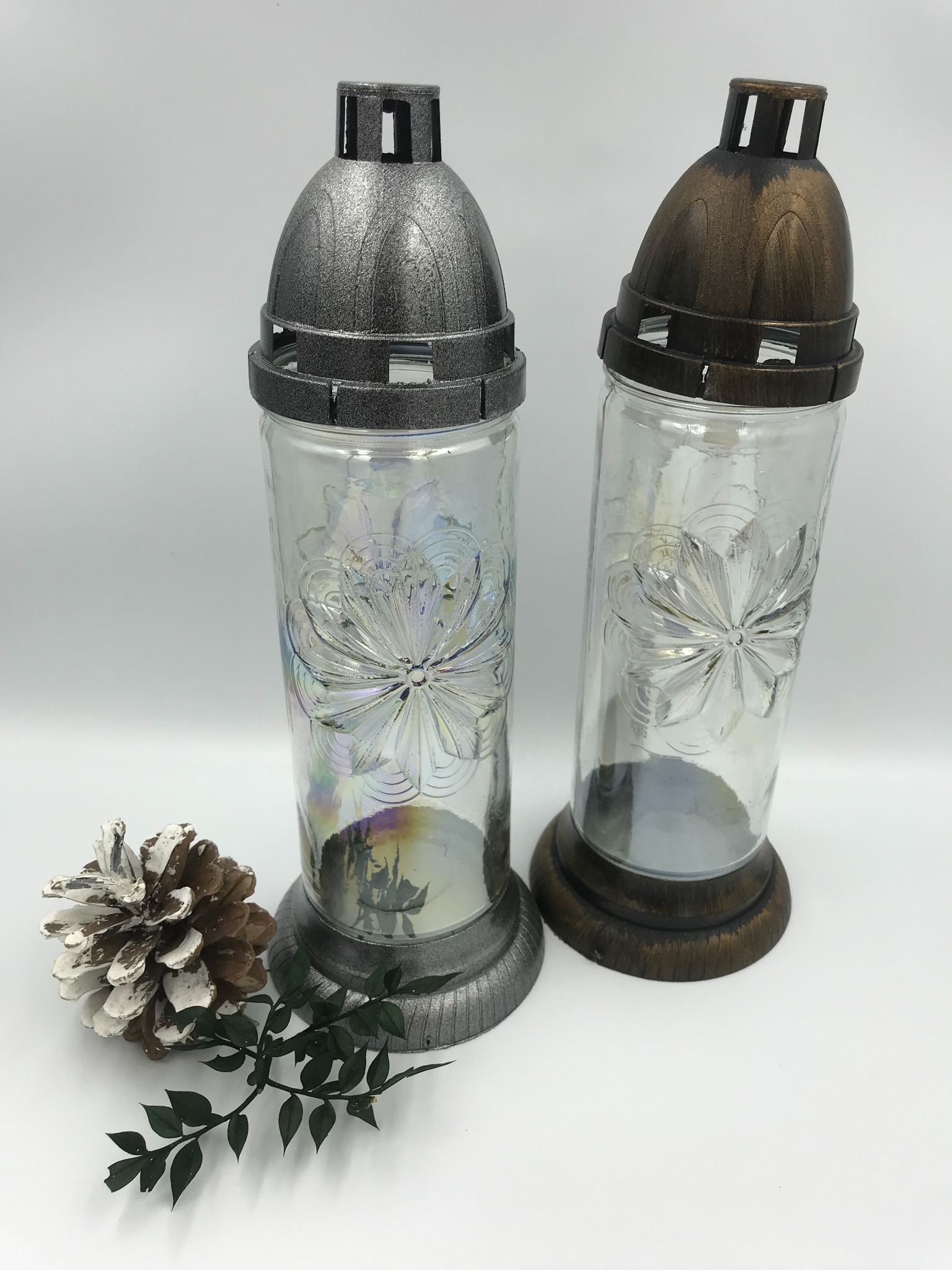 Dwa lampiony cmentarne ze szkła z wytłoczonym kwiatowym wzorem, jeden srebrny, drugi brązowy, leżą obok szyszki pomalowanej na biało i gałązki z zielonymi liśćmi na białym tle.