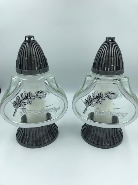 Dwa szklane znicze z metalowymi elementami i napisami 'Pamiętamy' i 'Kochamy', ustawione obok siebie na białym tle.