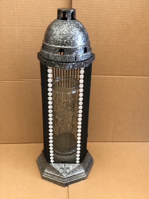 Czarno-srebrny lampion nagrobny z szklanym wkładem, ozdobiony białymi koralikami, na tle kartonowym.