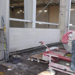 BUD-MAR Tetiana Martyniak - Wnętrze budowy: ściana z białych bloczków, rusztowanie i piła do cięcia. Widoczny kubeł i paleta. Surowy stan, przed wykończeniem.