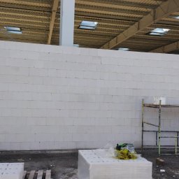 BUD-MAR Tetiana Martyniak - Wnętrze w trakcie budowy: ściana z białych bloczków, metalowa konstrukcja dachu z oknami, betonowe słupy i rusztowanie. Na podłodze materiały budowlane.