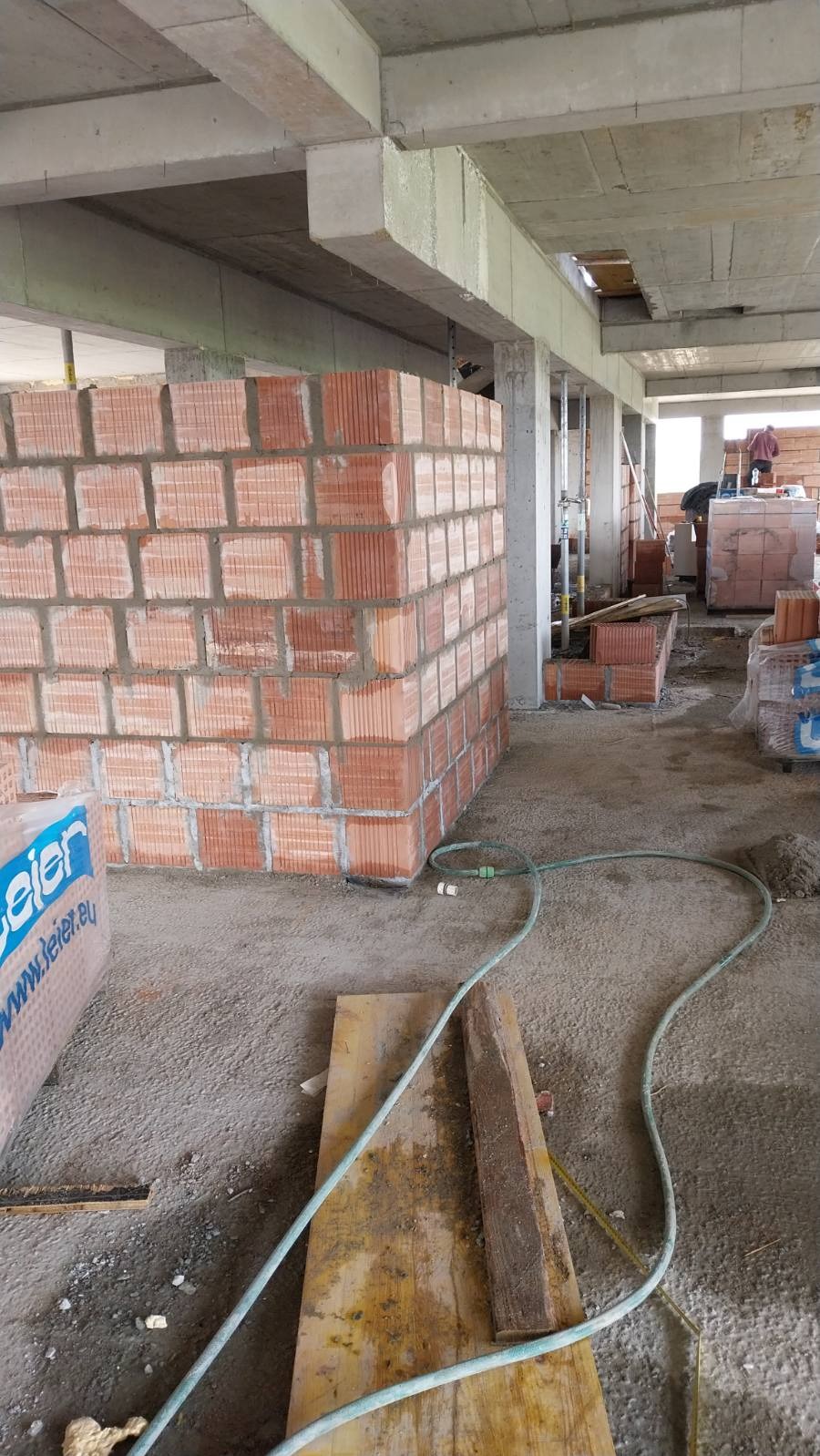 Wznoszenie ściany z czerwonej cegły na budowie. Widoczny fragment ściany, betonowy strop i słupy. W tle materiały budowlane i osoba na drabinie.