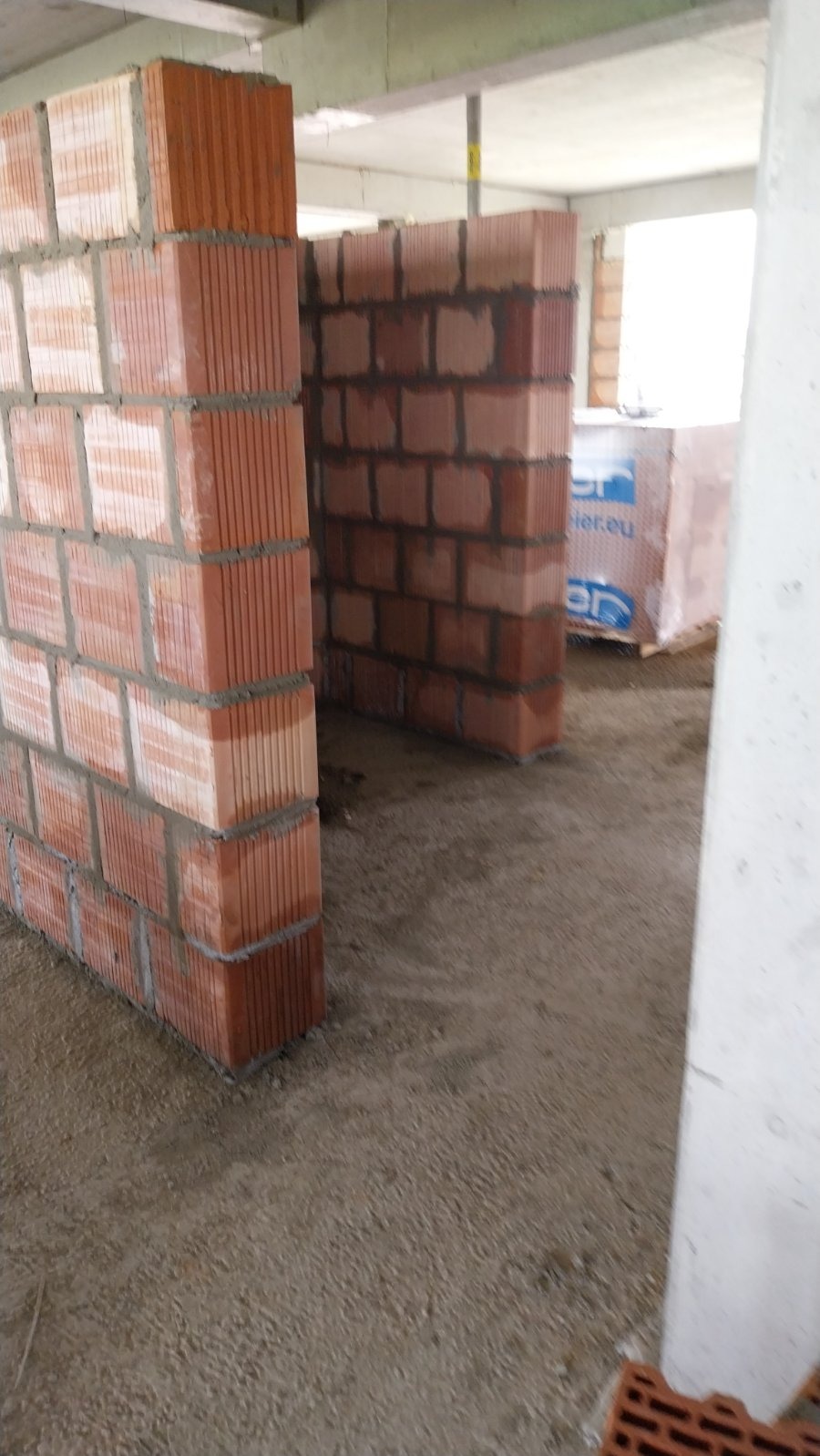 Wznoszenie ścian z czerwonej cegły ceramicznej, widoczne spoiny i palety z cegłami w tle. Etap budowy domu.