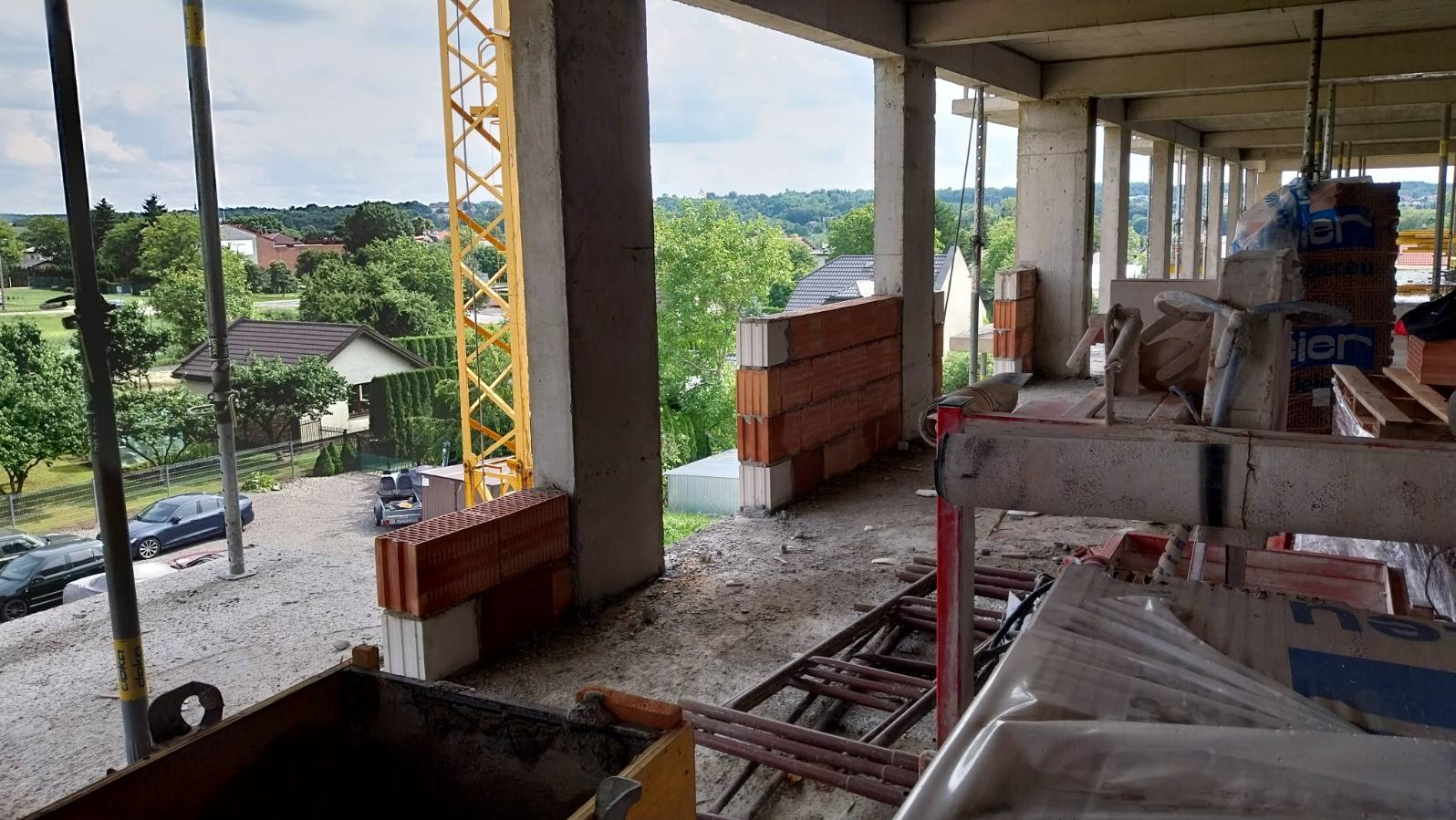 Wnętrze surowego budynku w trakcie murowania ścian. Widoczny dźwig budowlany, bloczki ceramiczne i narzędzia murarskie. W tle zabudowa jednorodzinna i zieleń.