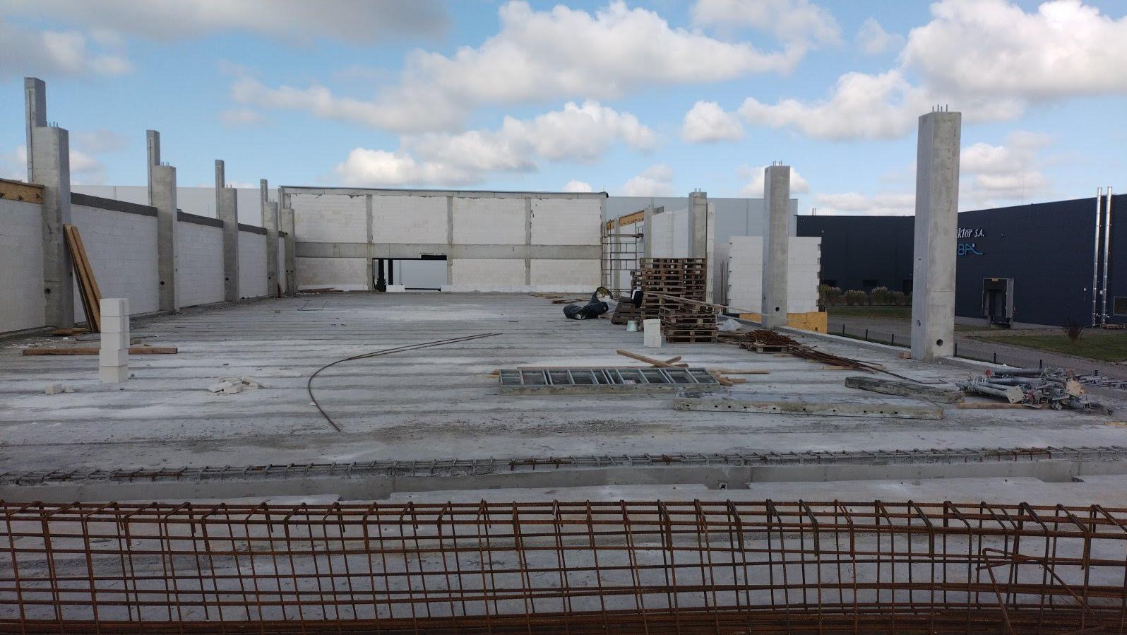 Budowa hali: betonowa płyta fundamentowa z wystającymi zbrojeniami, ściany z betonu komórkowego, słupy nośne. Widoczne materiały budowlane i palety.