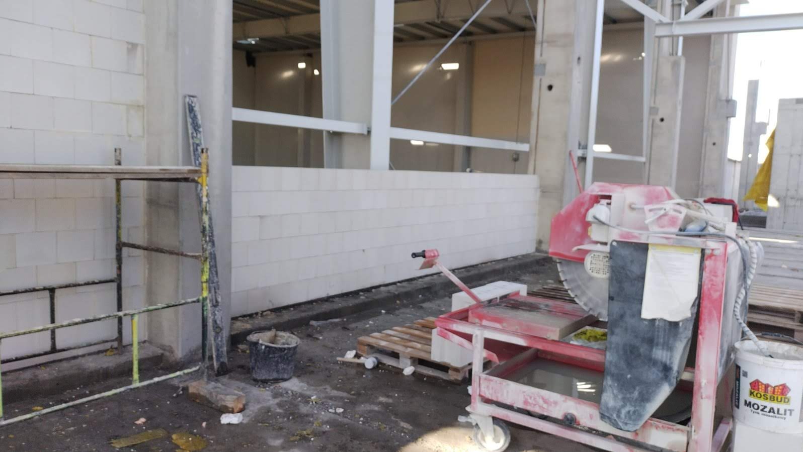 Wnętrze budowy: ściana z białych bloczków, rusztowanie i piła do cięcia. Widoczny kubeł i paleta. Surowy stan, przed wykończeniem.