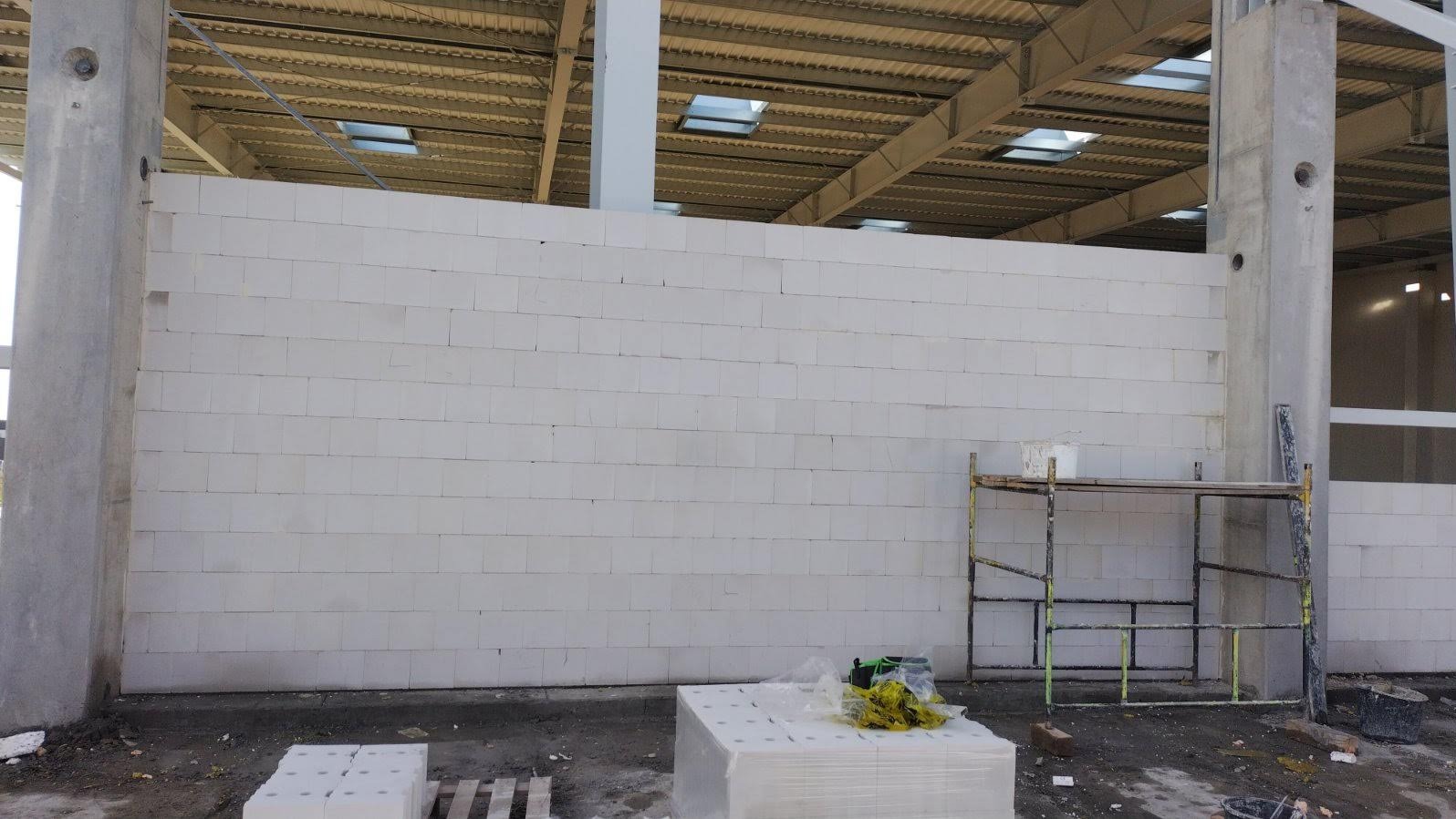 Wnętrze w trakcie budowy: ściana z białych bloczków, metalowa konstrukcja dachu z oknami, betonowe słupy i rusztowanie. Na podłodze materiały budowlane.