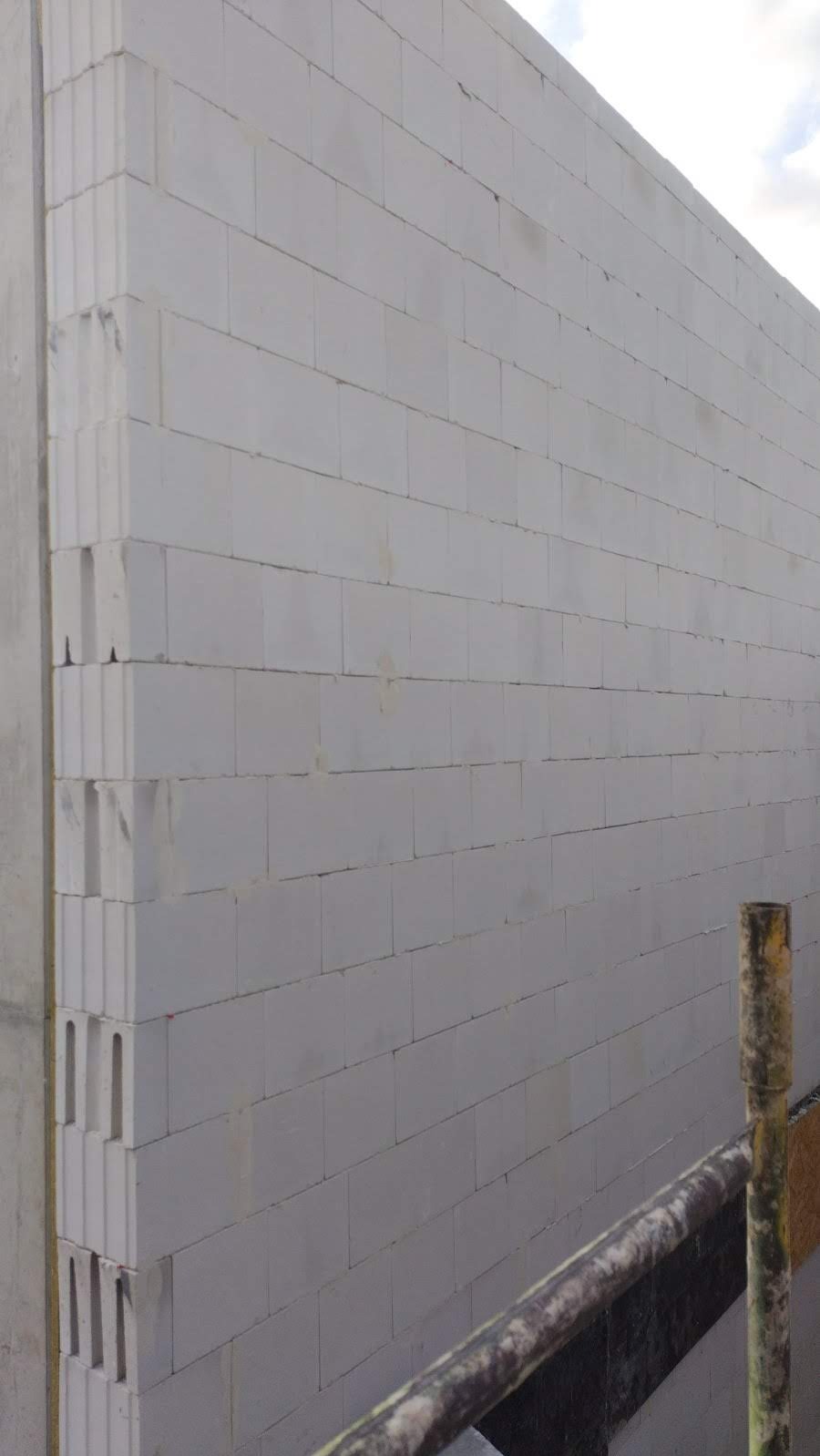 Ściana budynku w trakcie murowania z białych bloczków, widoczny narożnik i fragment rusztowania. Elementy konstrukcyjne w surowym stanie.