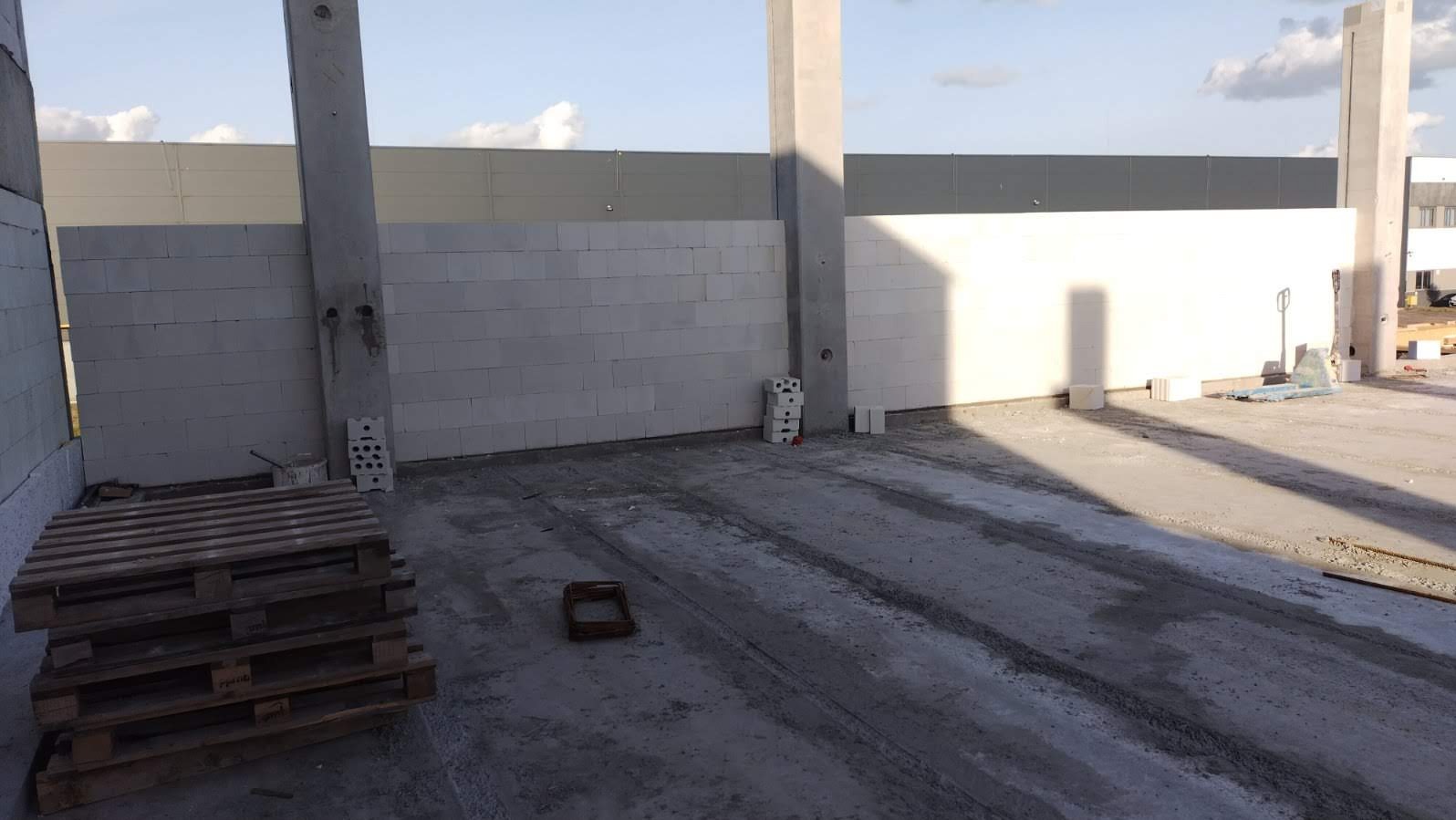 Wnętrze budynku w trakcie murowania ścian z białych bloczków. Widoczne betonowe kolumny, palety i ślady po maszynach na podłodze. Etap budowy w surowym stanie.