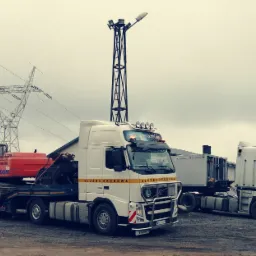 Ciężarówka Volvo z naczepą transportuje pomarańczową koparkę EX215; w tle linia wysokiego napięcia i druga ciężarówka.