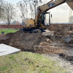 Żółta koparka CAT w trakcie prac ziemnych, wykop pod betonowy zbiornik z pokrywą, widoczna zielona maszyna na kołach w tle, ziemia rozkopana, w tle drzewa bez liści i czerwone ogrodzenie.