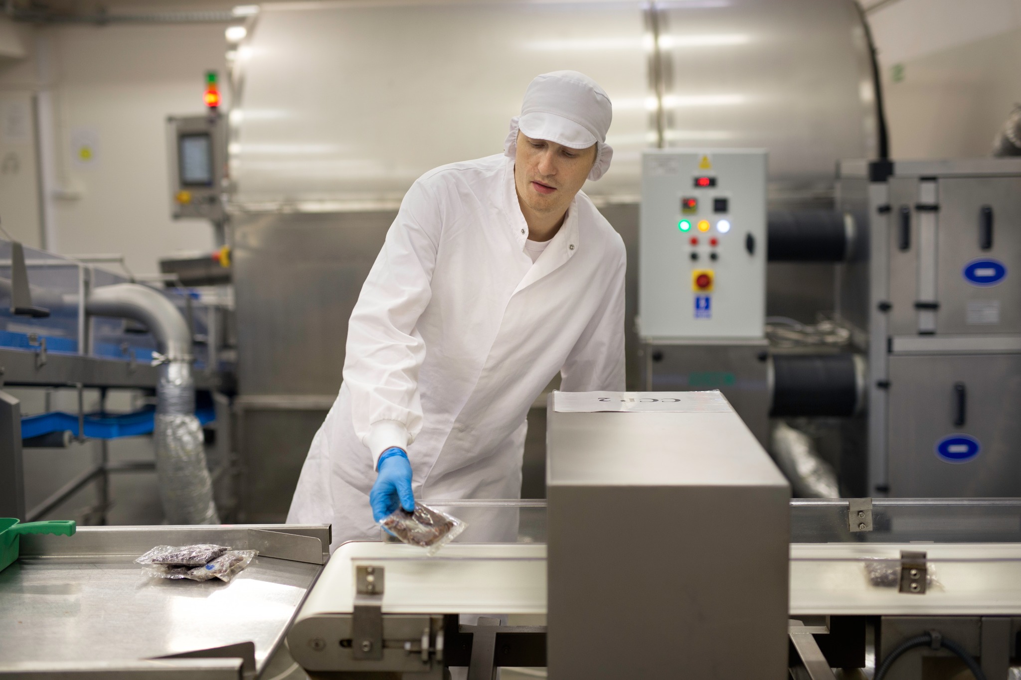 Pracownik w sterylnym ubraniu i rękawicach umieszcza zapakowane produkty spożywcze na linii produkcyjnej w zakładzie przetwórstwa, z widocznym panelem kontrolnym i metalowymi instalacjami w tle.