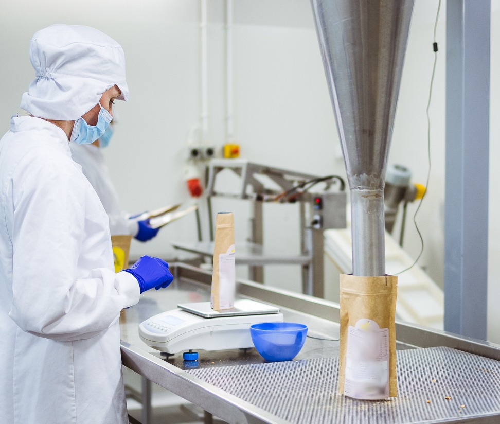 Pracownicy w sterylnych kombinezonach i rękawicach wypełniają brązowe papierowe torebki za pomocą metalowego lejka na linii produkcyjnej, z wagą i miską w pobliżu.