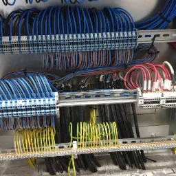 Rozdzielnica elektryczna z licznymi przewodami w różnych kolorach (niebieski, żółty, czarny, czerwony) podłączonymi do listew zaciskowych na szynach montażowych.