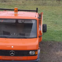 Pomarańczowa wywrotka Mercedes z włączonym pomarańczowym światłem ostrzegawczym na dachu, zaparkowana na żwirowej powierzchni, z zielonym trawnikiem w tle.