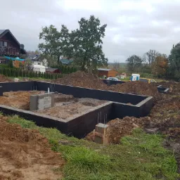 Wykopany i zabezpieczony fundament pod budowę domu jednorodzinnego, widoczne zbrojenia i izolacja, w tle dom z panelami słonecznymi i jesienny krajobraz.