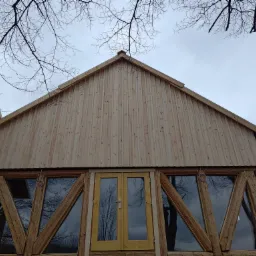 Surowy drewniany budynek z dwuspadowym dachem, widok z dołu, z widocznymi gałęziami drzew na tle pochmurnego nieba, okna z trójkątnymi podziałami szprosów.