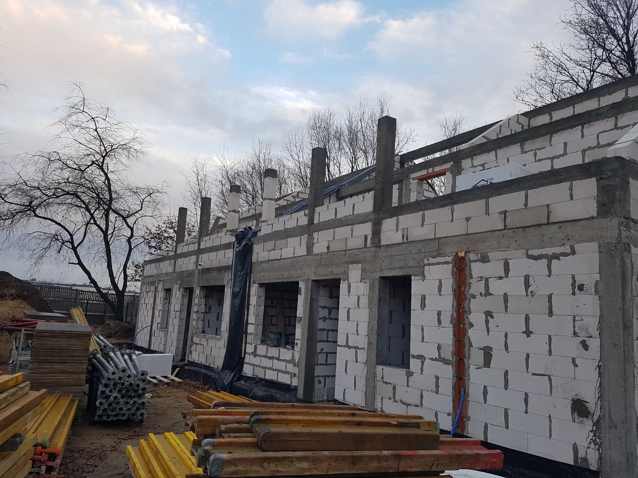 Budowa domu murowanego w Krakowie w trakcie realizacji, widoczne ściany z betonu komórkowego, drewniane stemple i materiały budowlane, pochmurne niebo w tle.