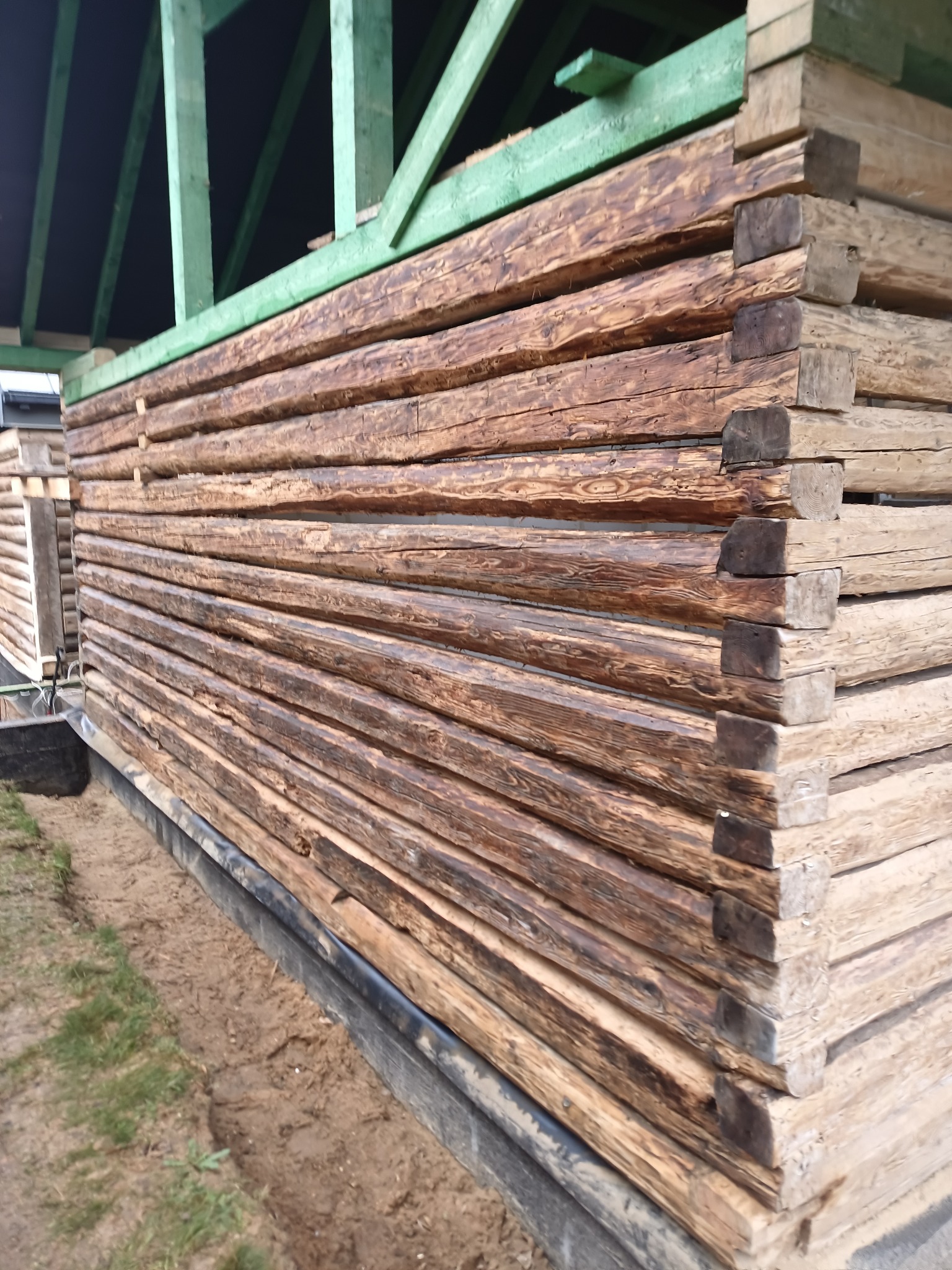 Fragment ściany zewnętrznej domu z bali, z widocznym połączeniem narożnym bali i zieloną konstrukcją dachu w tle.