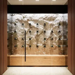 Nowoczesna piwniczka na wino za szklanymi drzwiami, z kamienną ścianą i drewnianą półką, oświetlona punktowo.