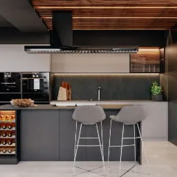 Nowoczesna kuchnia w odcieniach szarości z drewnianymi akcentami na suficie, wyspą z oświetloną półką na wino i dwoma szarymi hokerami, marmurowym backsplash'em i wbudowanymi urządzeniami.