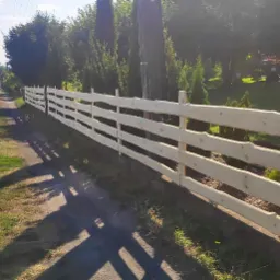 Długi, jasny, drewniany płot z surowego drewna o nieregularnych krawędziach, biegnący wzdłuż polnej drogi, z widocznymi cieniami i zielenią w tle.