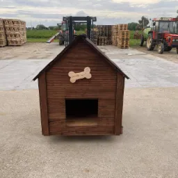 Drewniana buda dla psa z dekoracyjną kością na froncie, wykonana z ciemnego drewna, umieszczona na betonowym placu z paletami i maszynami rolniczymi w tle.