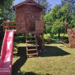 Drewniany plac zabaw w ogrodzie z domkiem na palach, czerwoną zjeżdżalnią, ścianką wspinaczkową i huśtawką na tle zieleni.