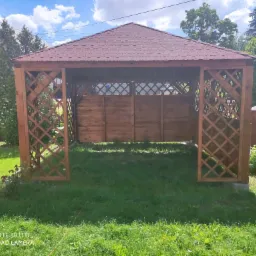 Drewniana pergola z brązowym dachem krytym gontem bitumicznym, częściowo osłonięta drewnianą kratą i panelami, na trawniku w ogrodzie.