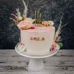 Tort urodzinowy dla Joli na 50 urodziny, zdobiony różami, trawami pampasowymi, złotymi świeczkami i napisem 'JOLA' na białym postumencie, na tle szarej ściany.