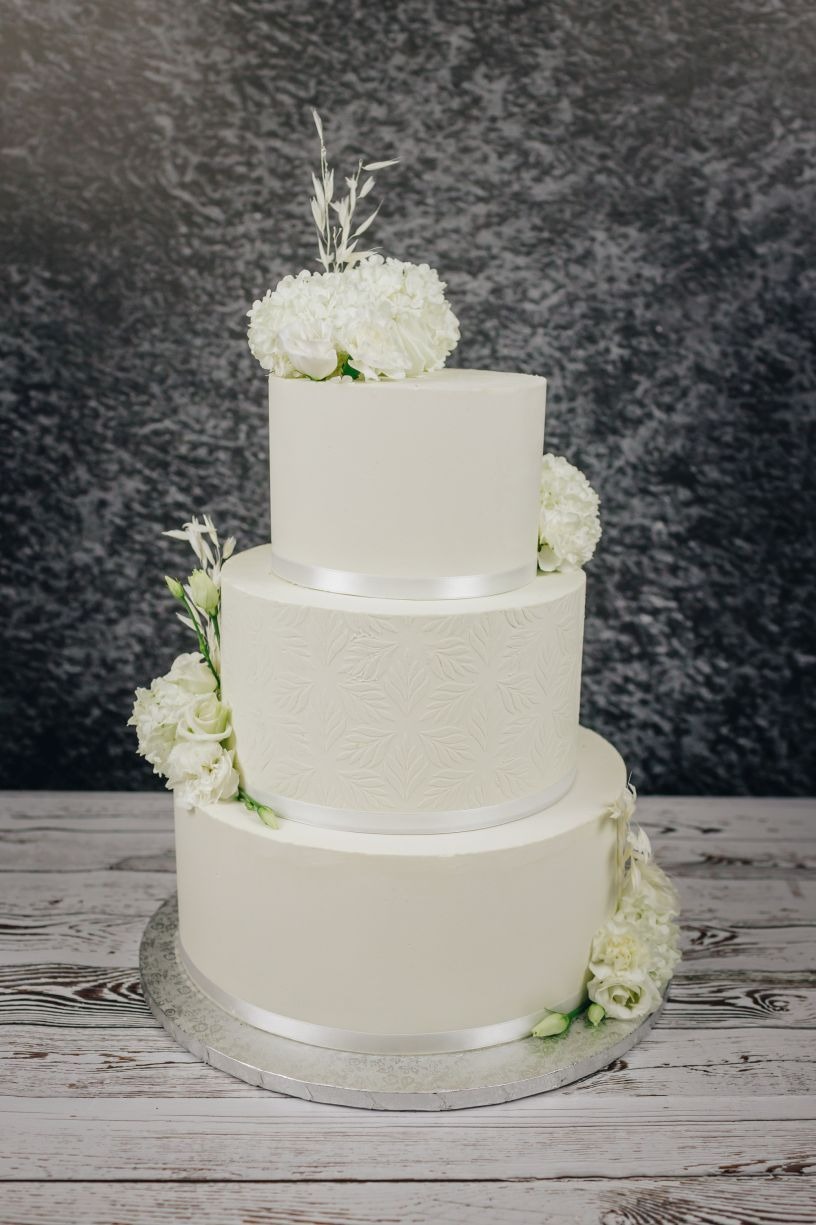 Trzypiętrowy tort ślubny w odcieniach bieli, zdobiony reliefem z motywem liści na środkowym piętrze, z satynowymi wstążkami, świeżymi białymi kwiatami i drobnymi źdźbłami trawy na szczycie...