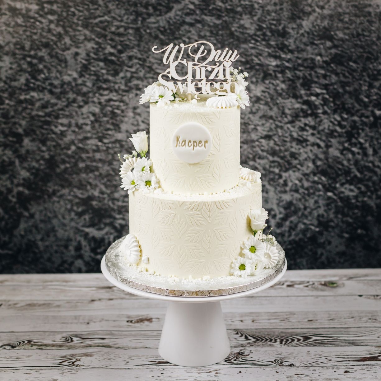 Dwupoziomowy tort w odcieniach bieli na Chrzest Święty, zdobiony motywem liści, świeżymi kwiatami i topperem 'W Dniu Chrztu Świętego' oraz imieniem 'Kacper', ustawiony na białej paterze...