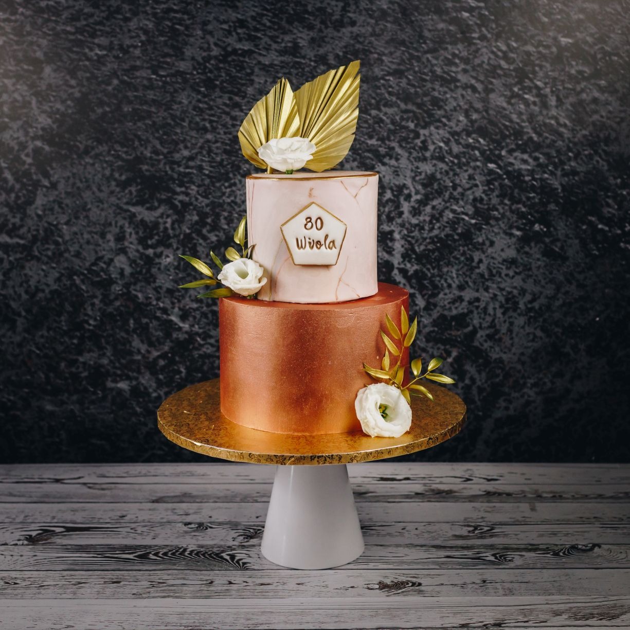 Dwupiętrowy tort urodzinowy w odcieniach różu i miedzi, zdobiony złotymi liśćmi, białymi kwiatami i napisem '80 Wiola' w geometrycznej ramce, umieszczony na złotym talerzu na białej nodze.