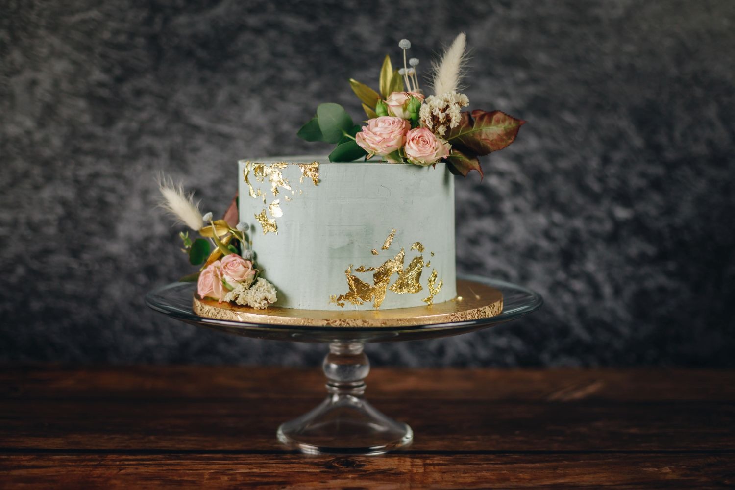 Tort w pastelowym odcieniu zieleni, zdobiony płatkami złota i kompozycją z różowych róż oraz suszonych traw na szklanej paterze.