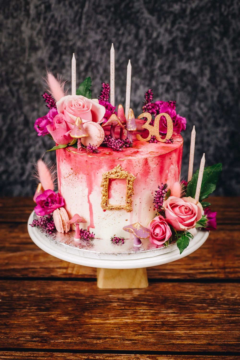 Tort urodzinowy w odcieniach różu i złota, zdobiony kwiatami, makaronikami i świeczkami, stojący na białym paterze na drewnianym stole, z dekoracyjną ramką na boku.